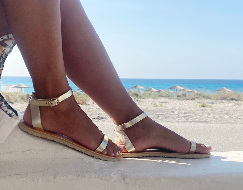 May include: A pair of metallic gold flat sandals featuring a thin strap across the toes and an adjustable ankle strap with a small gold-toned buckle. The sandals have a flat, light-colored sole and a simple, open design that secures around the ankle.