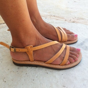 May include: A pair of tan leather sandals featuring multiple thin straps across the top of the foot, a flat dark brown sole with visible white stitching, and an adjustable ankle strap secured with a small metallic buckle on the side.