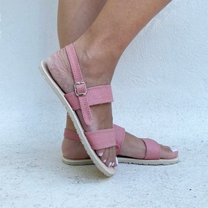 May include: A pair of dusty rose sandals featuring two horizontal straps across the top of the foot, a back heel strap with a silver-toned metal buckle closure, and a flat, light-colored sole with visible stitching along the edges.