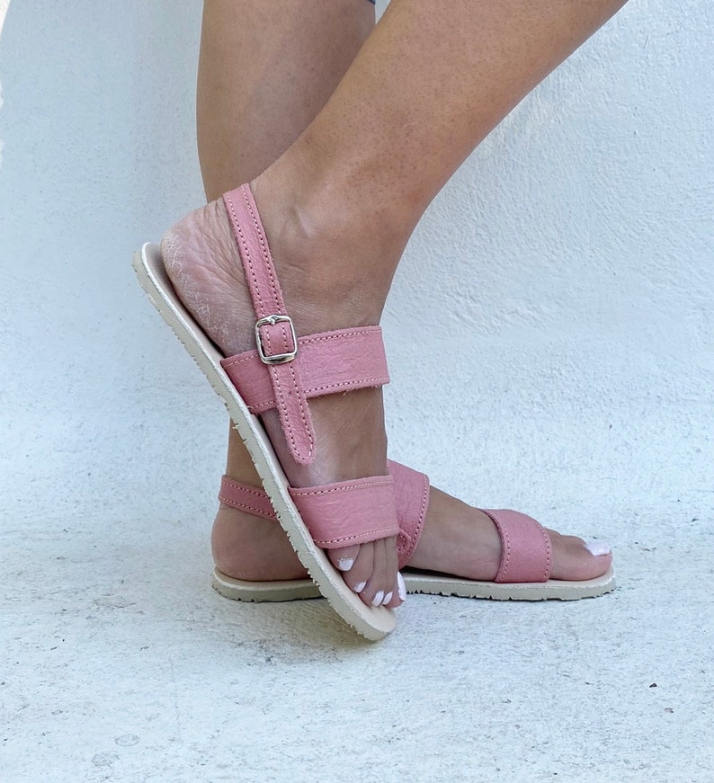 May include: A pair of dusty rose sandals featuring two horizontal straps across the top of the foot, a back heel strap with a silver-toned metal buckle closure, and a flat, light-colored sole with visible stitching along the edges.