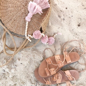 Op de afbeelding: Een ronde, gevlochten tas van beige stro met een grote oudroze kwast en een snoer van lichte houten kralen met drie oudroze pompons. Naast de tas liggen twee beige leren sandalen in gladiatorenstijl met lange, dunne veters om te strikken.