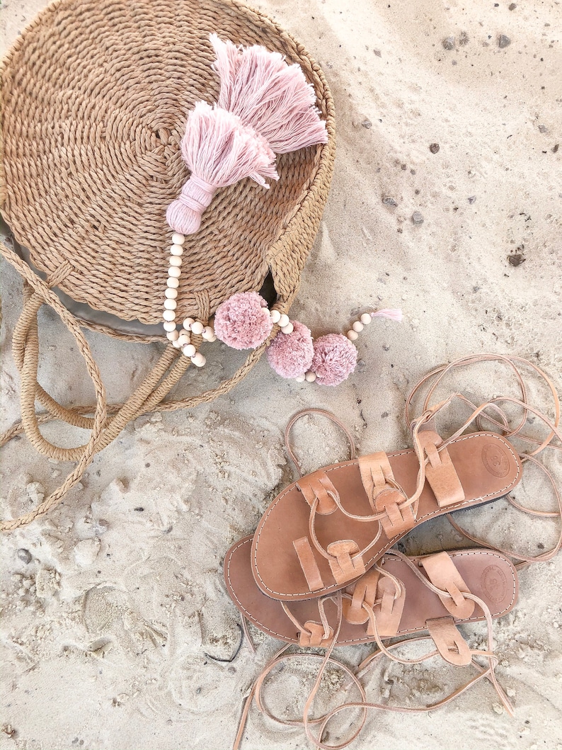 Op de afbeelding: Een ronde, gevlochten tas van beige stro met een grote oudroze kwast en een snoer van lichte houten kralen met drie oudroze pompons. Naast de tas liggen twee beige leren sandalen in gladiatorenstijl met lange, dunne veters om te strikken.