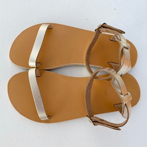 May include: A pair of flat sandals featuring metallic gold straps across the toes and around the ankles. The footbeds are tan with the number 36 printed on the heel area. The ankle straps include small gold-toned metal buckles for adjustment.