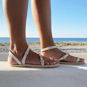 May include: White flat sandals featuring a thin ankle strap with a gold-toned metal buckle and a toe loop. The upper strap is decorated with small, circular, textured gold-toned embellishments arranged in a row along the top of the foot.