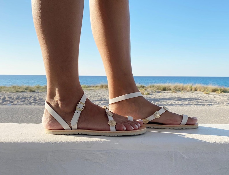 May include: White flat sandals featuring a thin ankle strap with a gold-toned metal buckle and a toe loop. The upper strap is decorated with small, circular, textured gold-toned embellishments arranged in a row along the top of the foot.