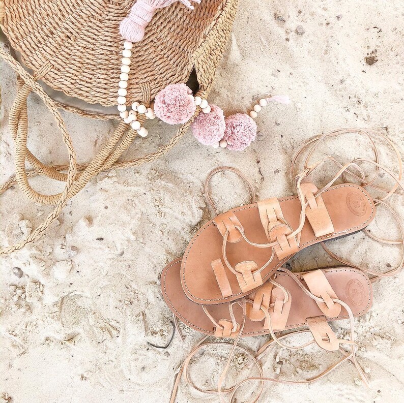 Op de afbeelding: Een paar beige leren vetersandalen met platte zolen en gevlochten bandjes, geplaatst naast een ronde tas van gevlochten stro, versierd met een snoer van lichte houten kralen en drie zachte, oudroze pompons aan het handvat.