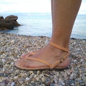 May include: A pair of brown leather sandals with a buckle closure. The sandals are worn by a person standing on a beach with small pebbles.