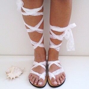 May include: White lace sandals with straps that wrap around the ankles and legs. The sandals have a simple design and are perfect for a beach wedding or other special occasion.