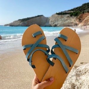 Hand holding a pair of handmade Greek sandals with the blue sea in the background, perfect for summer vacations and beach walks."