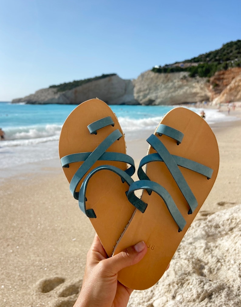 Hand holding a pair of handmade Greek sandals with the blue sea in the background, perfect for summer vacations and beach walks."