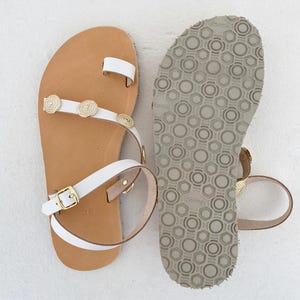 May include: A pair of white leather sandals featuring a toe loop, gold-toned circular metal embellishments on the strap, and an adjustable ankle strap with a gold buckle. The tan footbed is marked with the size 36, and the sole has a grey tread.