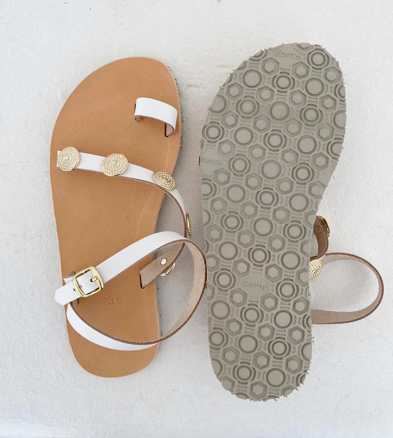 May include: A pair of white leather sandals featuring a toe loop, gold-toned circular metal embellishments on the strap, and an adjustable ankle strap with a gold buckle. The tan footbed is marked with the size 36, and the sole has a grey tread.