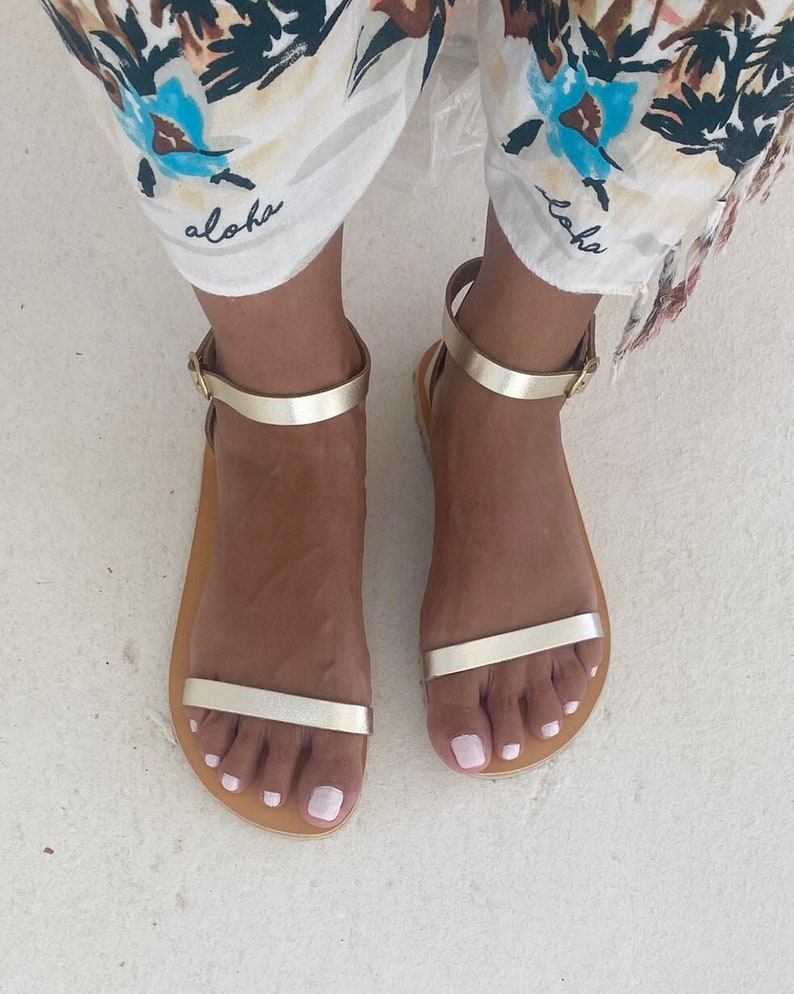 May include: A pair of flat sandals featuring metallic gold straps across the toes and around the ankles with a simple buckle closure. The sandals have a tan-colored footbed and a minimalist design, providing a clean look for warm weather footwear.