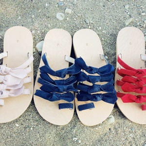 May include: Three pairs of sandals with multiple bows on each strap. The sandals are beige with pink, navy blue, and red bows.