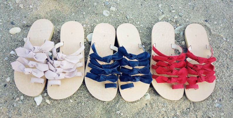 May include: Three pairs of sandals with multiple bows on each strap. The sandals are beige with pink, navy blue, and red bows.
