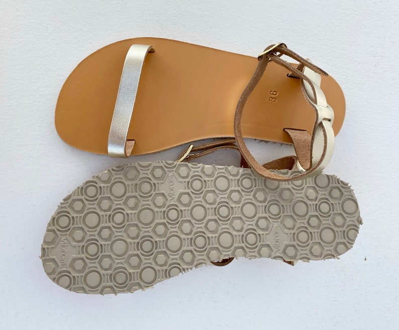 May include: A pair of flat sandals featuring a metallic silver toe strap and a tan leather footbed with the number 36 stamped on the heel. The sandals have a beige rubber Vibram outsole with a geometric circular and hexagonal tread pattern.