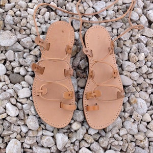 Op de afbeelding: Een paar beige leren gladiatorsandalen met een teenlus, meerdere horizontale bandjes en lange veters om rond de enkel te wikkelen. Op het voetbed van elke sandaal staat het cijfer 9 gestempeld, wat de schoenmaat aangeeft.