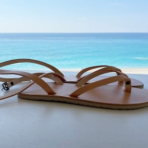 May include: Two pairs of tan leather sandals with adjustable straps and buckles. The sandals are on a white surface with a view of the ocean in the background.