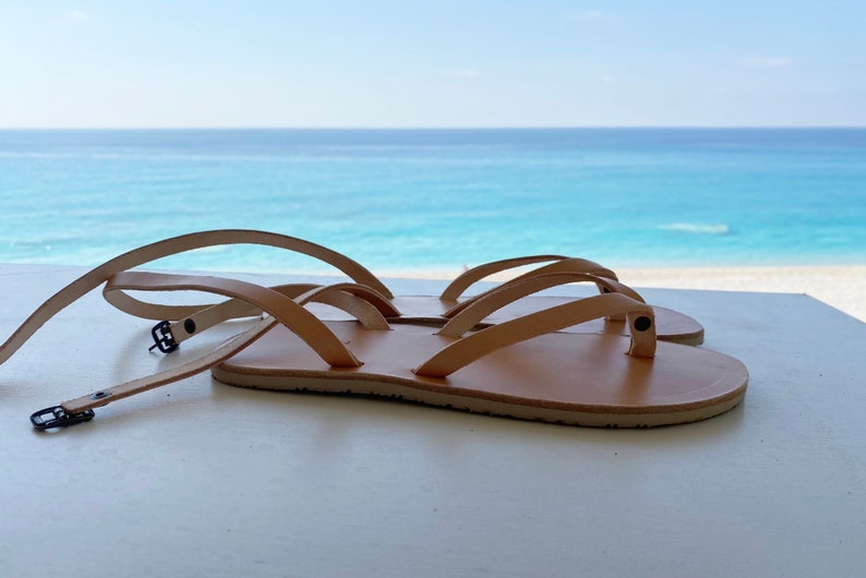 May include: Two pairs of tan leather sandals with adjustable straps and buckles. The sandals are on a white surface with a view of the ocean in the background.