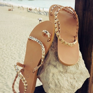 K&ouml;nnte beinhalten: Ein Paar braune Ledersandalen mit goldenen geflochtenen Riemen. Die Sandalen liegen auf einem Stein mit einem Strand im Hintergrund.