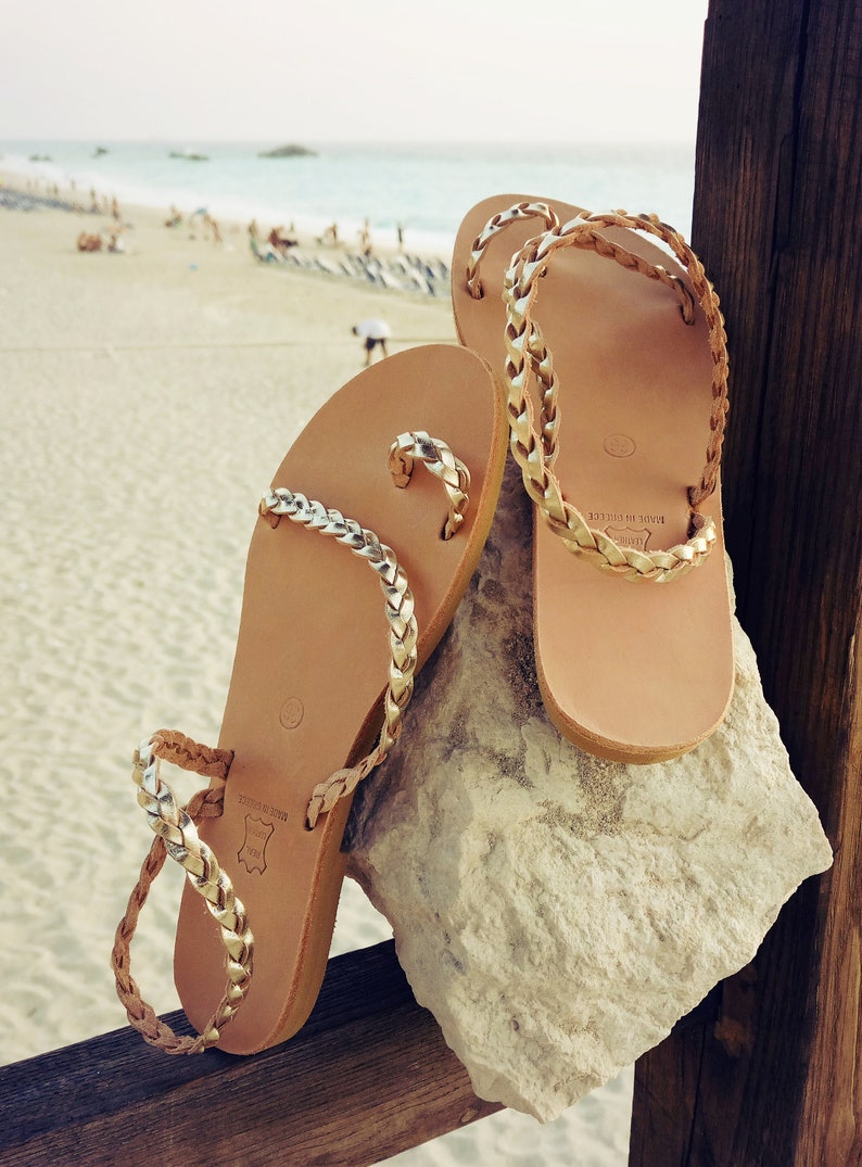 K&ouml;nnte beinhalten: Ein Paar braune Ledersandalen mit goldenen geflochtenen Riemen. Die Sandalen liegen auf einem Stein mit einem Strand im Hintergrund.