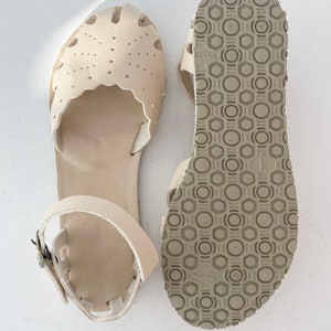 May include: A pair of cream-colored leather sandals featuring a scalloped edge, perforated detailing on the toe box, and an adjustable ankle strap with a metal buckle. The underside of the sole displays a textured, geometric Vibram tread pattern.