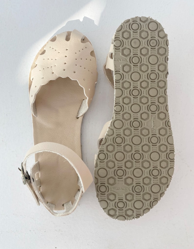 May include: A pair of cream-colored leather sandals featuring a scalloped edge, perforated detailing on the toe box, and an adjustable ankle strap with a metal buckle. The underside of the sole displays a textured, geometric Vibram tread pattern.