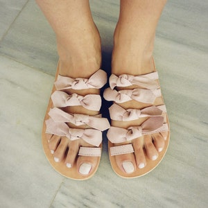 May include: A pair of blush pink sandals with multiple bow straps. The sandals are made of suede and have a leather sole.