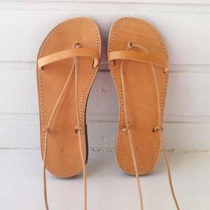 May include: A pair of tan leather sandals with laces that tie up the legs. The sandals have a simple design and are made of high-quality leather.
