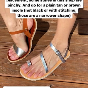 May include: A pair of flat sandals featuring metallic silver straps and a tan leather insole. One sandal has a toe loop and an ankle strap with a metal buckle, while the other features a wide metallic silver band across the forefoot and a brown ankle strap.