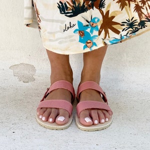 May include: A pair of dusty rose sandals with two horizontal straps and a buckle closure, featuring a light-colored textured sole. The sandals are worn beneath a cream-colored skirt with a tropical print that includes the word aloha in script.