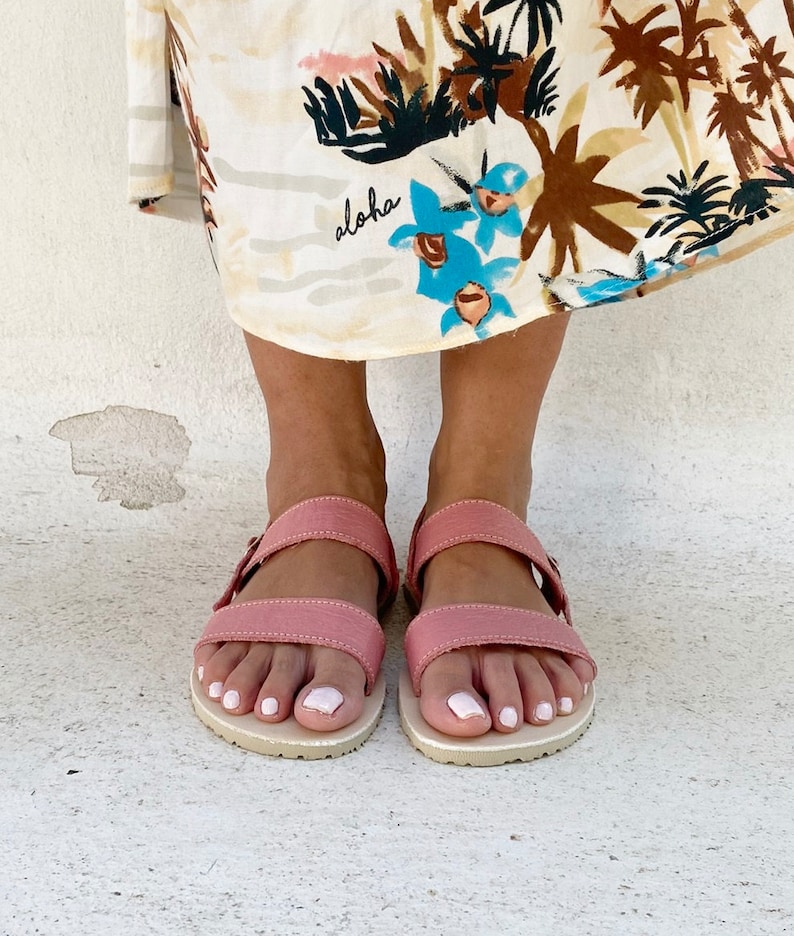 May include: A pair of dusty rose sandals with two horizontal straps and a buckle closure, featuring a light-colored textured sole. The sandals are worn beneath a cream-colored skirt with a tropical print that includes the word aloha in script.