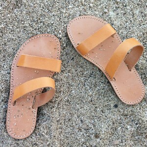 A pair of brown leather sandals with two straps. The sandals are on a sandy surface.