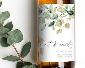 May include: A white wine bottle with a green and gold floral design label. The label reads "aunt and uncle" with a heart symbol and "EST. AUGUST 2024 BABY THOMPSON IS ON THE WAY!"