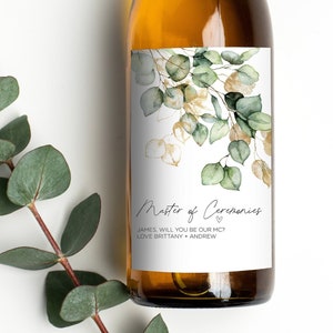 May include: A white wine bottle with a green and gold floral design label. The label reads "Master of Ceremonies" with the text "James, will you be our MC? Love Brittany + Andrew".