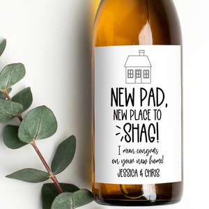 May include: A wine bottle with a gold-coloured top and a white label. The label features a house illustration and the text "NEW PAD, NEW PLACE TO SHAG! I mean congrats on your new home! JESSICA & CHRIS."