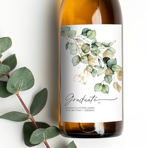May include: A white wine bottle with a green and gold eucalyptus leaf design label. The label reads "Graduate" with the text "Congratulations James Love Brittany + Andrew".