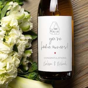 Könnte beinhalten: Eine Flasche Weißwein mit einem personalisierten Etikett, auf dem "You're home owners! Congratulations Chelsea & Michael" mit einem kleinen roten Herzen steht. Das Etikett zeigt auch eine einfache Strichzeichnung eines Hauses.