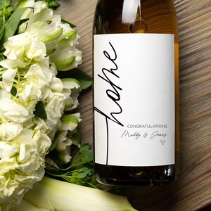 May include: A white wine bottle with a white label that says "Cheers" in black cursive writing. The label also says "Congratulations" and "Molly & Josh" in black text. The bottle is sitting on a wooden surface.