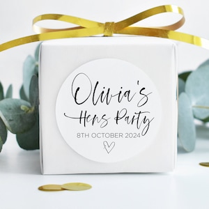 May include: A white gift box with a gold ribbon tied around it. A white round sticker with black text reads "Olivia's Hens Party 8th October 2024" with a small heart symbol.