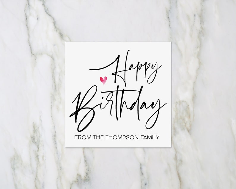 May include: A white card with black calligraphy that reads "Happy Birthday" with a small pink heart above the word "Birthday". The bottom of the card reads "From the Thompson Family".
