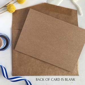 May include: A brown card with a blank back. The card is sitting on a white surface with a blue and white ribbon, a brown envelope, and yellow flowers.