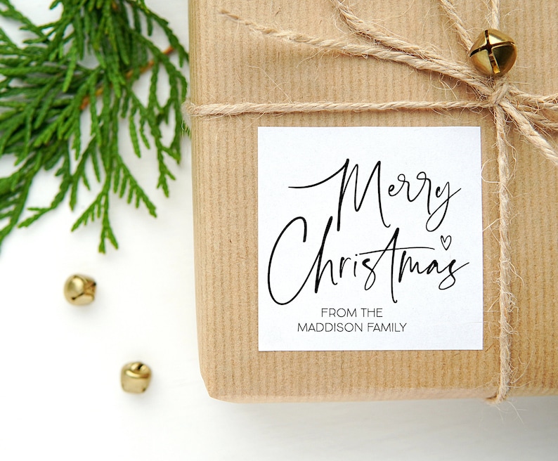 May include: A white gift tag with black calligraphy that reads "Merry Christmas" with a small heart symbol. The tag is attached to a brown paper gift box with twine and a gold bell.