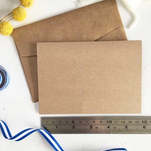 May include: Two brown envelopes, one slightly larger than the other, are stacked on a white surface. A ruler is placed below the envelopes, and a blue and white striped ribbon is curled on the surface.