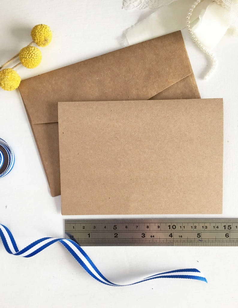 May include: Two brown envelopes, one slightly larger than the other, are stacked on a white surface. A ruler is placed below the envelopes, and a blue and white striped ribbon is curled on the surface.