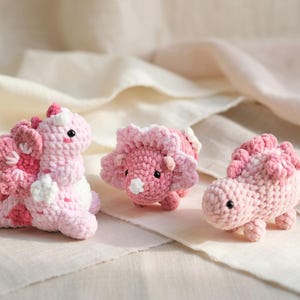 Könnte beinhalten: Drei handgefertigte Häkeldinosaurier-Spielzeuge in Rosa- und Weißtönen. Die Dinosaurier haben detaillierte Merkmale, darunter Hörner, Stacheln und kleine schwarze Augen. Die Spielzeuge werden auf einem cremefarbenen Stoff präsentiert, der die weiche Textur des Garns zeigt.