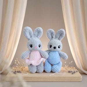Könnte beinhalten: Zwei handgefertigte Häkelhasen-Puppen. Einer trägt ein rosa Kleid, der andere ein blaues Outfit. Beide haben graue Körper, lange Ohren und schwarze Augen. Sie stehen auf einer Holzoberfläche vor einem neutralen Hintergrund.