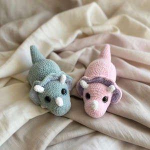 Könnte beinhalten: Zwei gehäkelte Triceratops-Dinosaurier. Einer ist hellblau mit weißen und hellvioletten Akzenten, der andere ist rosa mit weißen und hellvioletten Akzenten. Beide haben schwarze Augen und liegen auf einem beigen Stoff.