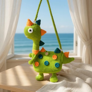 Könnte beinhalten: Handgefertigte grüne Häkeltasche in Dinosaurierform mit orangefarbenen, türkisfarbenen und gelben Akzenten. Die Tasche hat einen langen grünen Riemen und Knöpfe. Der Dinosaurier hat ein freundliches Gesicht.