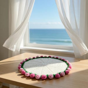 Könnte beinhalten: Ein handgefertigter, cremefarbener Häkel-Tischset mit grünem Rand und rosa Pompons. Die runde Matte liegt auf einem Holztisch in der Nähe eines Fensters mit weißen Vorhängen und Blick auf das Meer.